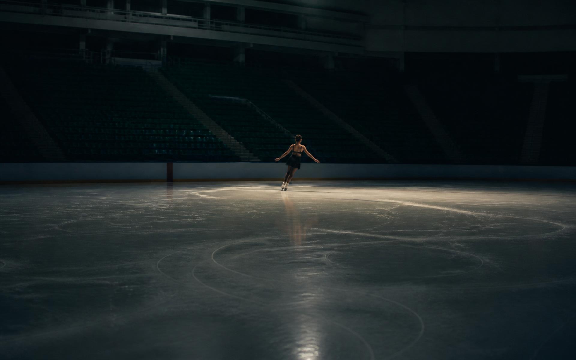 A single figure skater under quiet arena light, tracing curves across freshly resurfaced ice